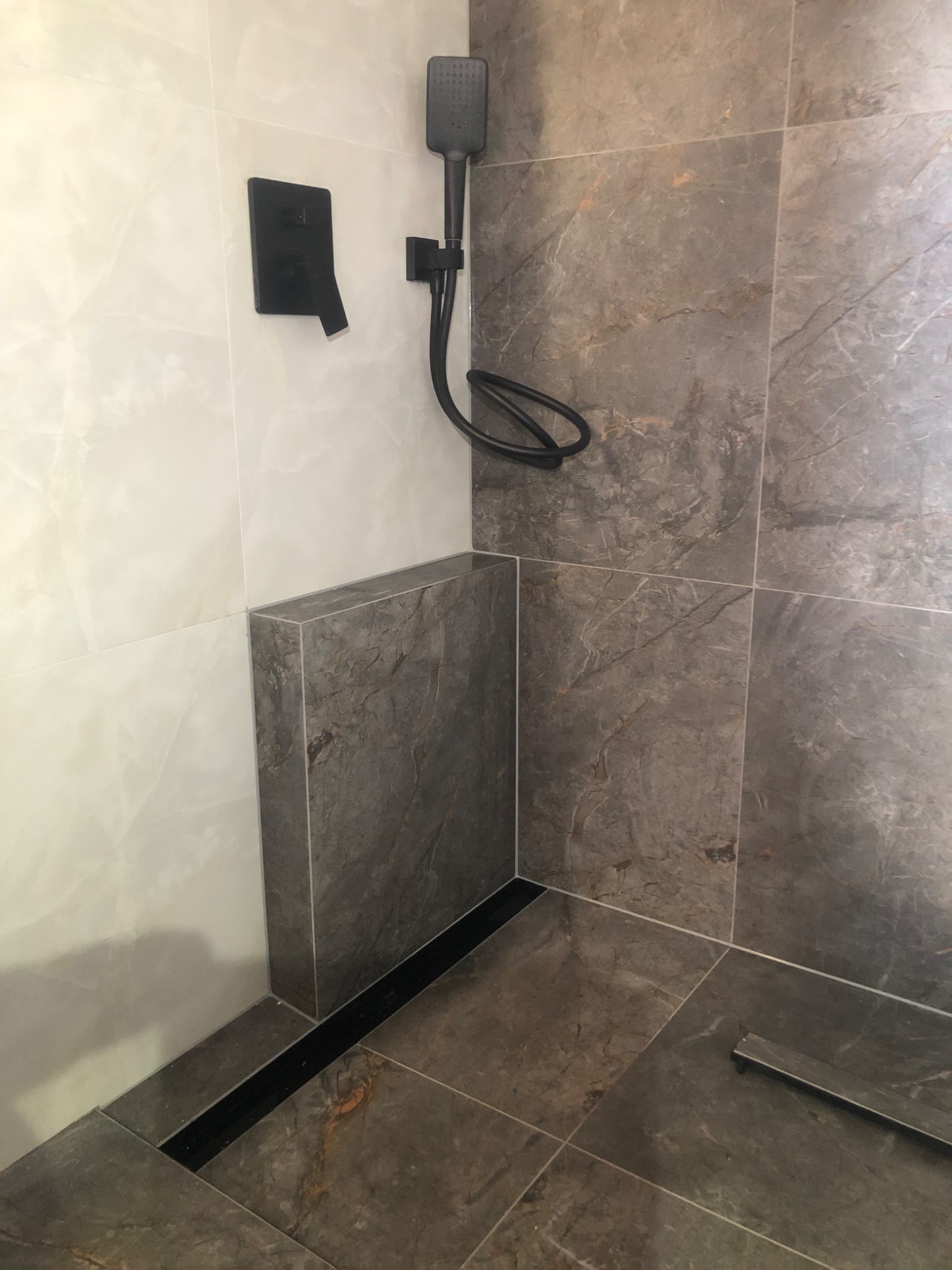 Nowoczesna kabina prysznicowa z szarymi płytkami imitującymi kamień, czarną armaturą i odpływem liniowym. Kontrastujące białe płytki po lewej stronie.