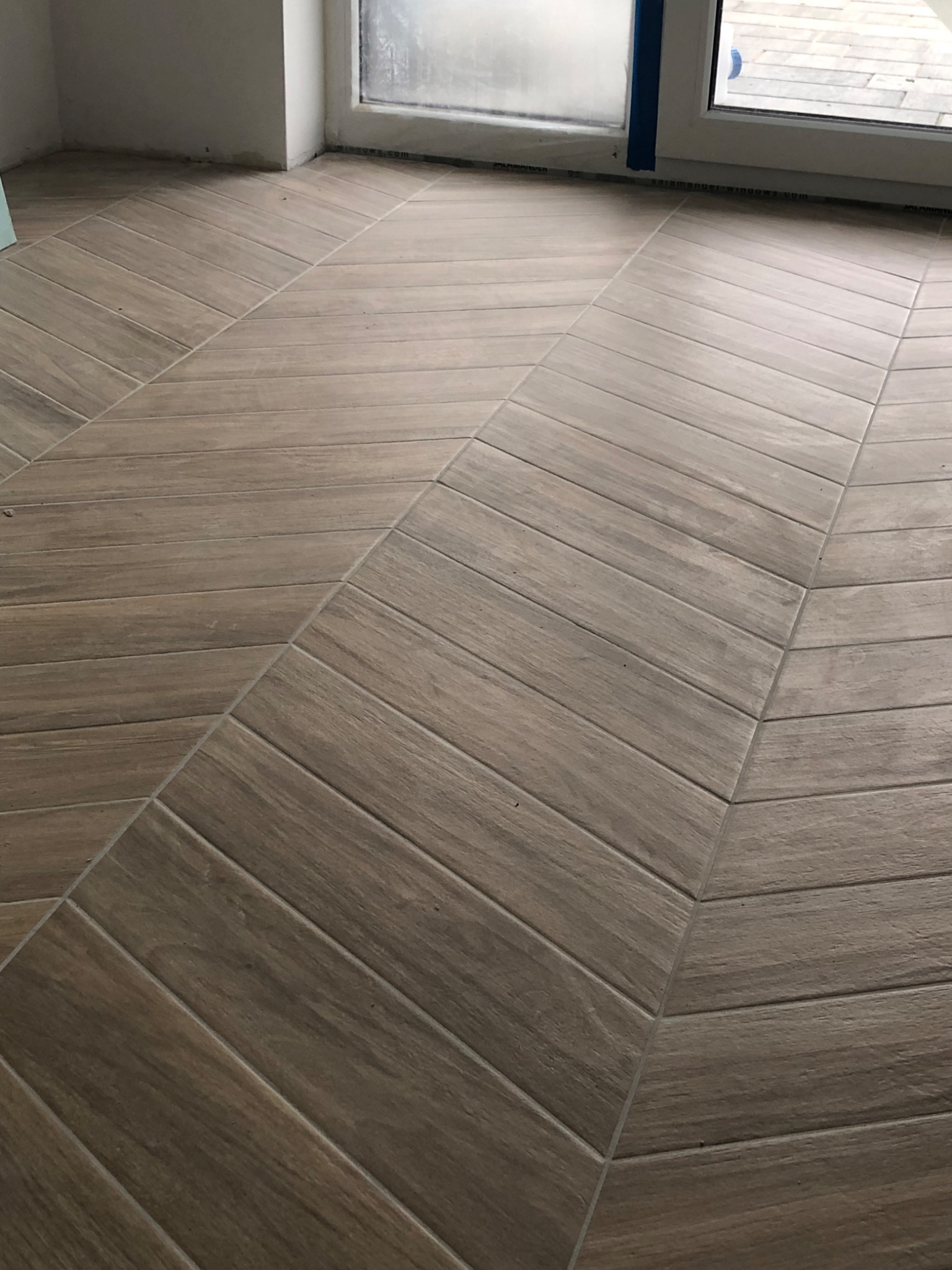 Ukończona podłoga z płytek imitujących drewno, ułożonych w jodełkę, przy oknie zabezpieczonym taśmą malarską. Ciemniejsze fugi podkreślają wzór.