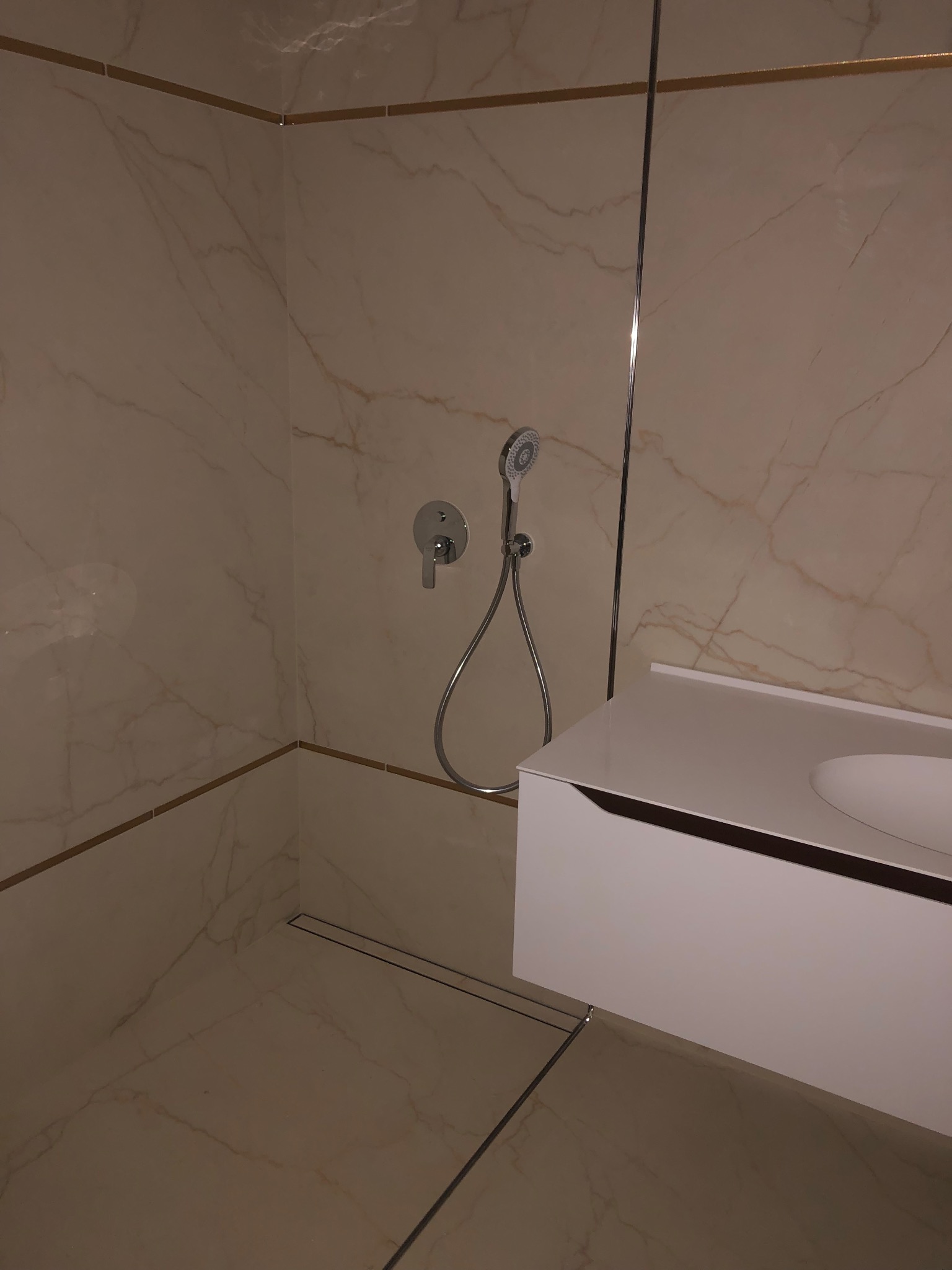 Wykończona łazienka z beżowymi płytkami imitującymi marmur, chromowaną armaturą i białą szafką pod umywalką. Widoczny odpływ liniowy w podłodze.