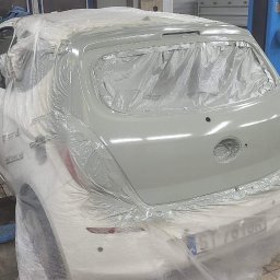 MAGNUSON CARS S.C. - Biały samochód na podnośniku w warsztacie, częściowo oklejony folią ochronną i pokryty świeżym podkładem w kolorze szarym, przygotowany do lakierowania.