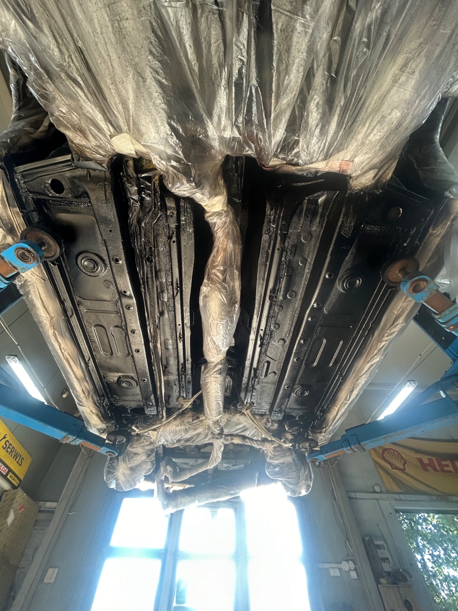 Podwozie samochodu po nałożeniu czarnej powłoki antykorozyjnej, zabezpieczone folią na podnośniku w warsztacie.