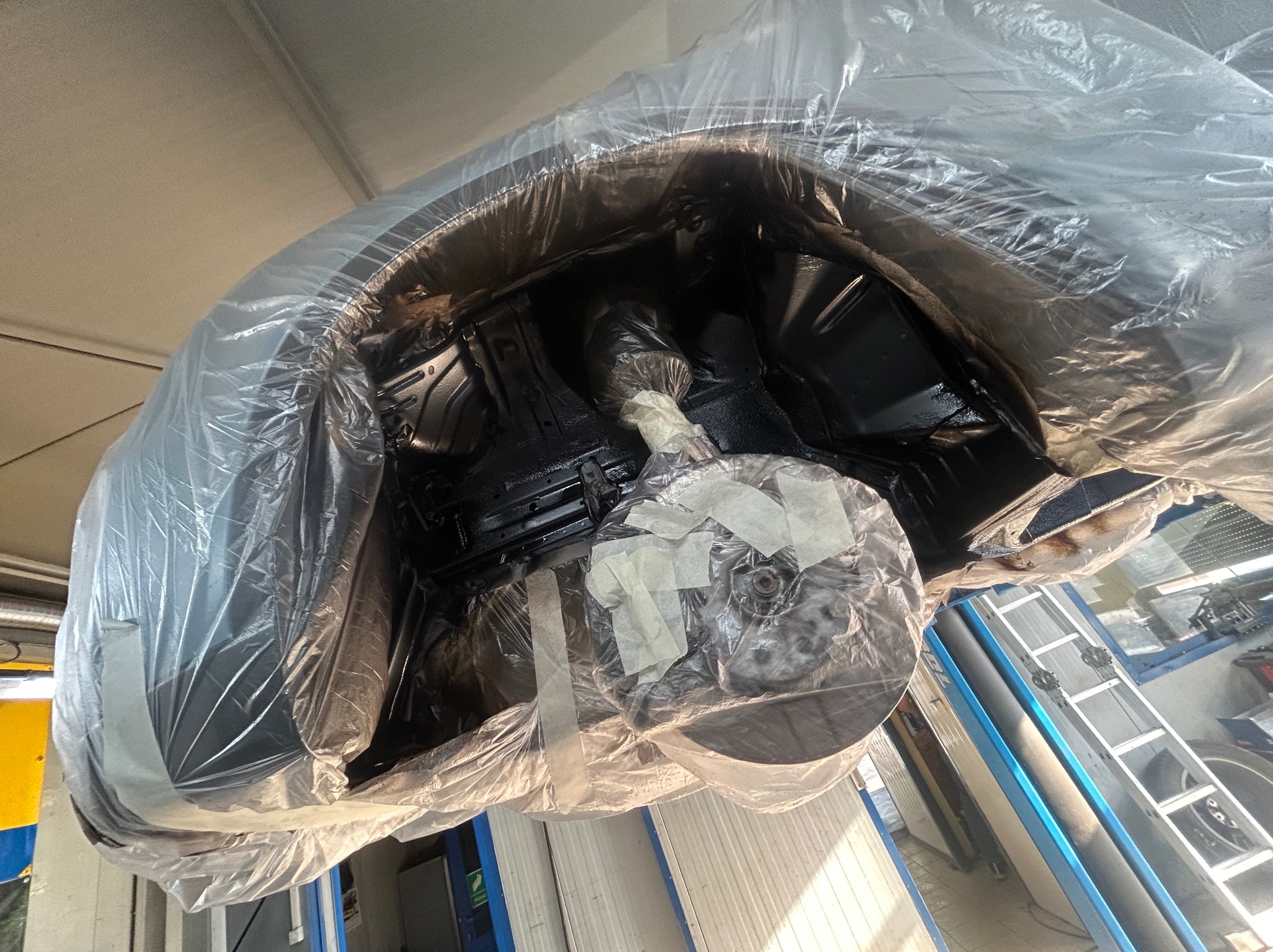 Nadwozie samochodu zabezpieczone folią i taśmą malarską przed lakierowaniem, widoczne elementy zawieszenia i podwozia w czarnym kolorze, warsztat samochodowy w tle.