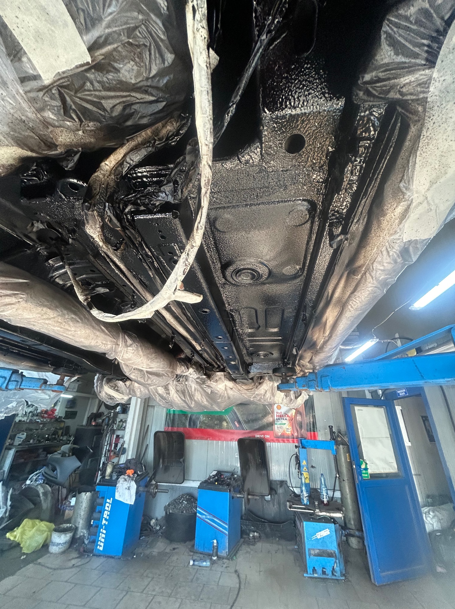 Podwozie samochodu po zabezpieczeniu antykorozyjnym, widoczne elementy zawieszenia i rury osłonięte folią, podnośnik warsztatowy w tle.