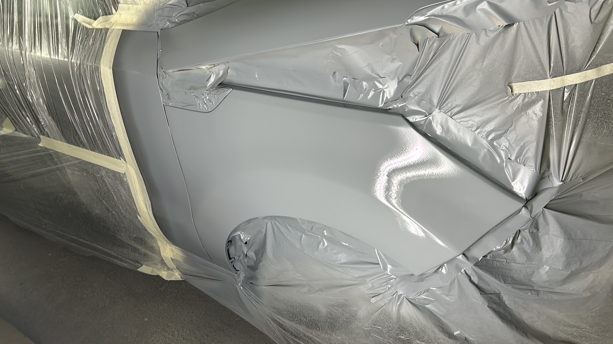 Fragment karoserii samochodu po świeżym lakierowaniu na szaro, zabezpieczony folią i taśmą malarską.