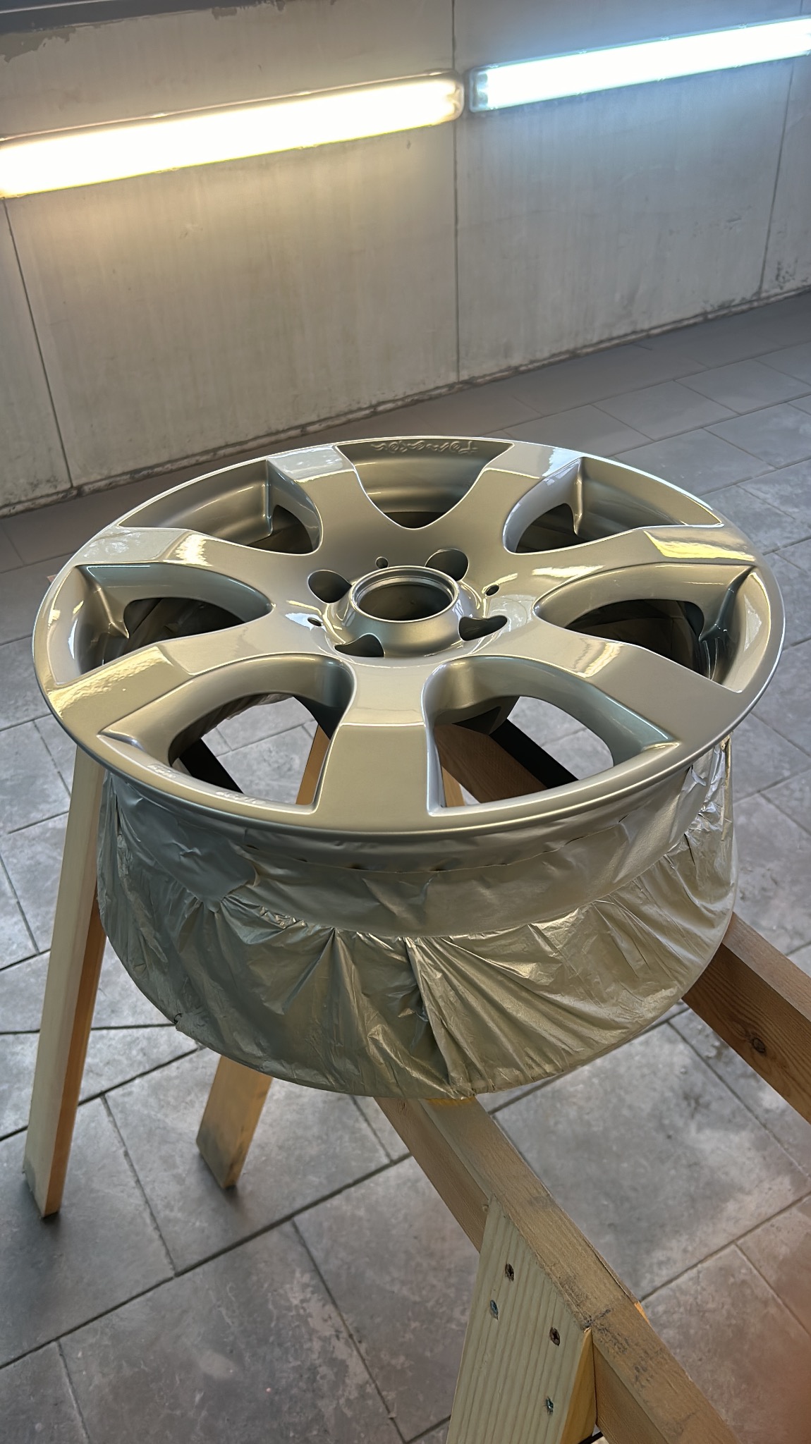 Pomalowana felga aluminiowa na drewnianym stojaku, zabezpieczona folią malarską, w warsztacie z szarą podłogą i jasnym oświetleniem.