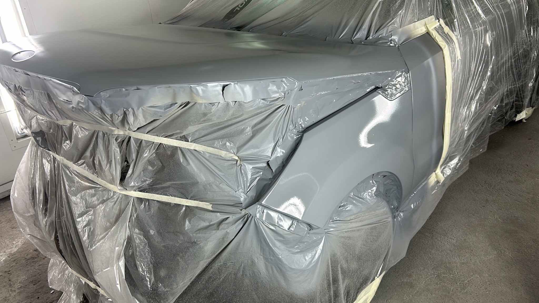 Samochód w trakcie lakierowania, zabezpieczony folią i taśmą malarską, świeżo pomalowany na szaro, widoczne refleksy światła na lakierze.
