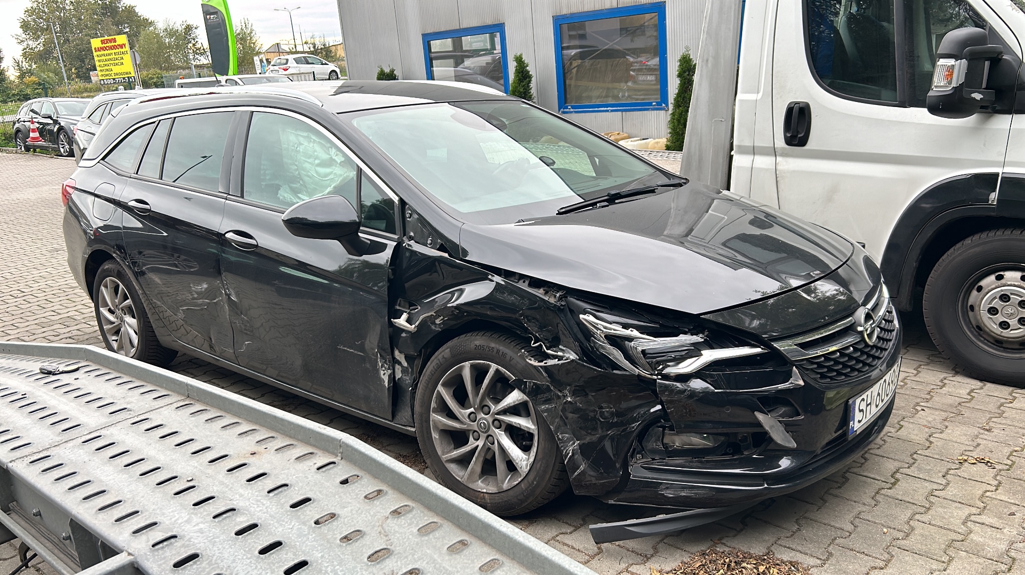 Uszkodzony przód czarnego kombi Opel Astra na lawecie, widoczne wgniecenia i pęknięcia karoserii oraz uszkodzone reflektory, obok biały samochód dostawczy.