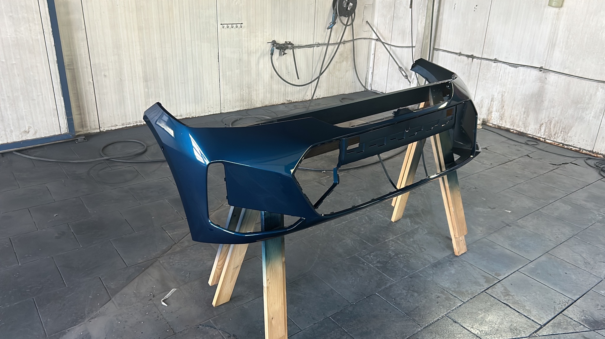 Pomalowany na ciemnoniebiesko zderzak samochodowy, umieszczony na drewnianych kozłach w warsztacie, widoczne przewody i narzędzia lakiernicze na ścianie w tle.