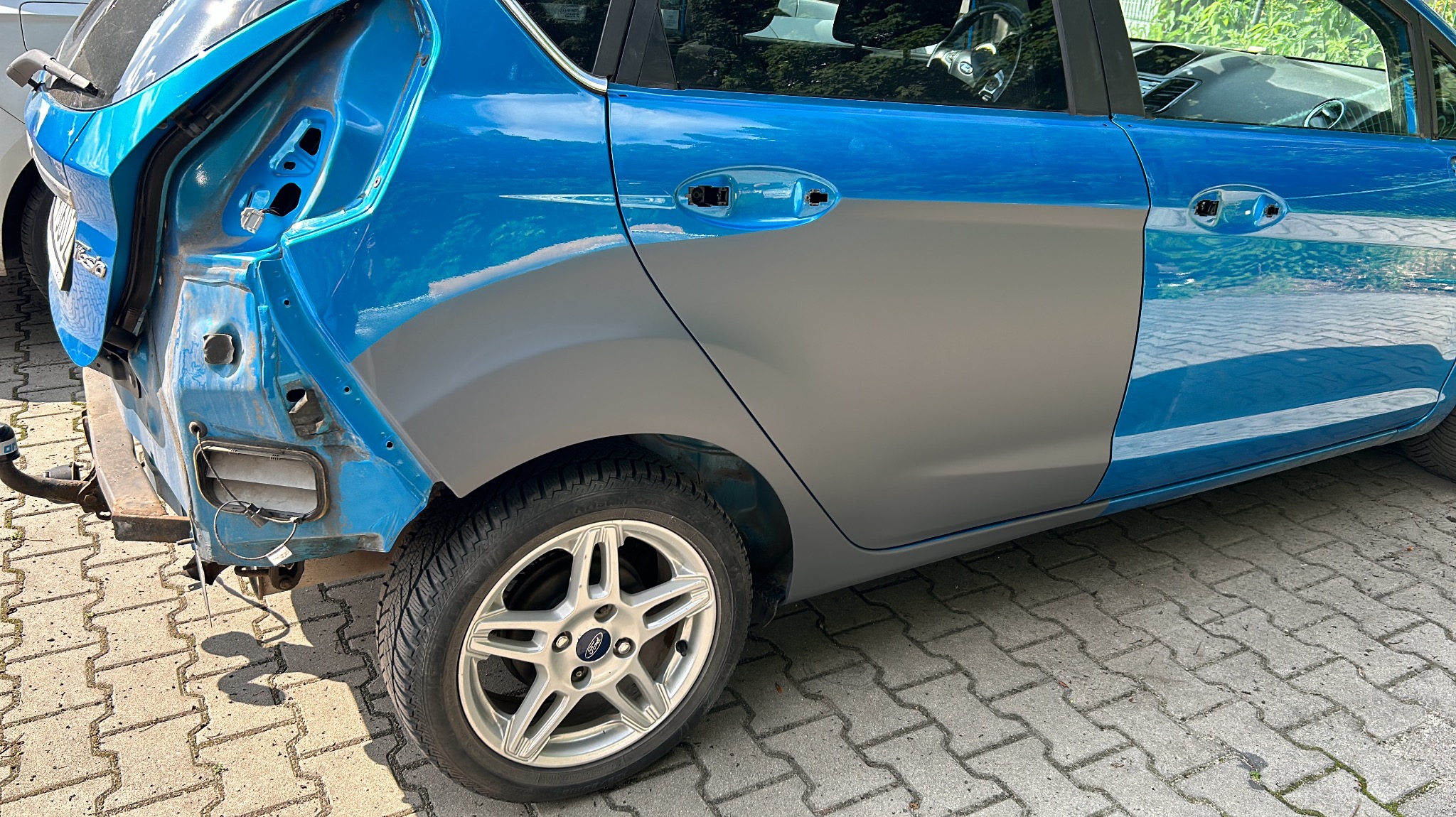 Niebieski samochód po wypadku, z uszkodzonym tyłem i przygotowany do lakierowania, z szarym podkładem na drzwiach i błotniku.