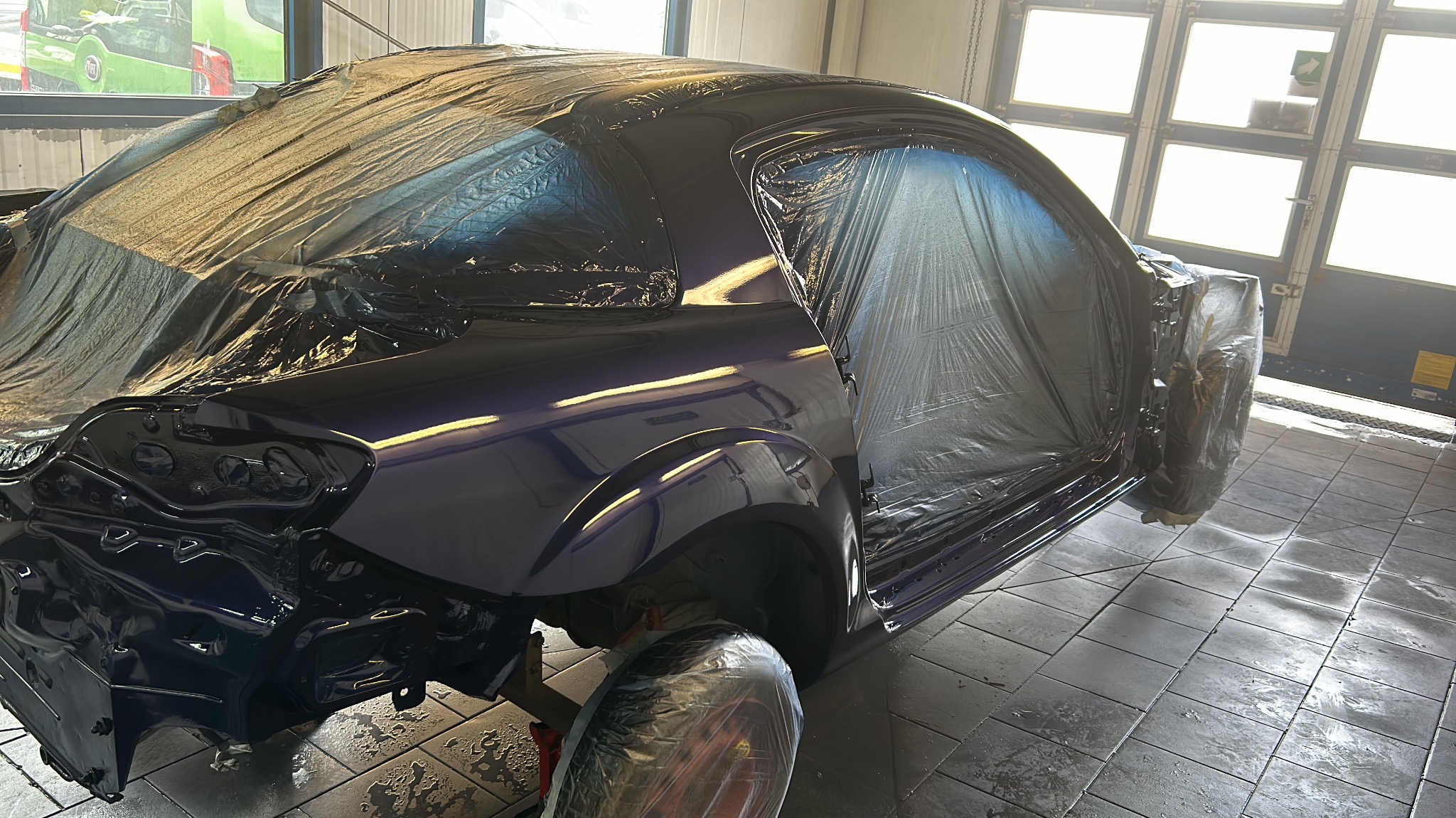 Przygotowanie samochodu do lakierowania: fioletowe auto owinięte folią ochronną w warsztacie blacharsko-lakierniczym, widoczne elementy karoserii i podwozia.