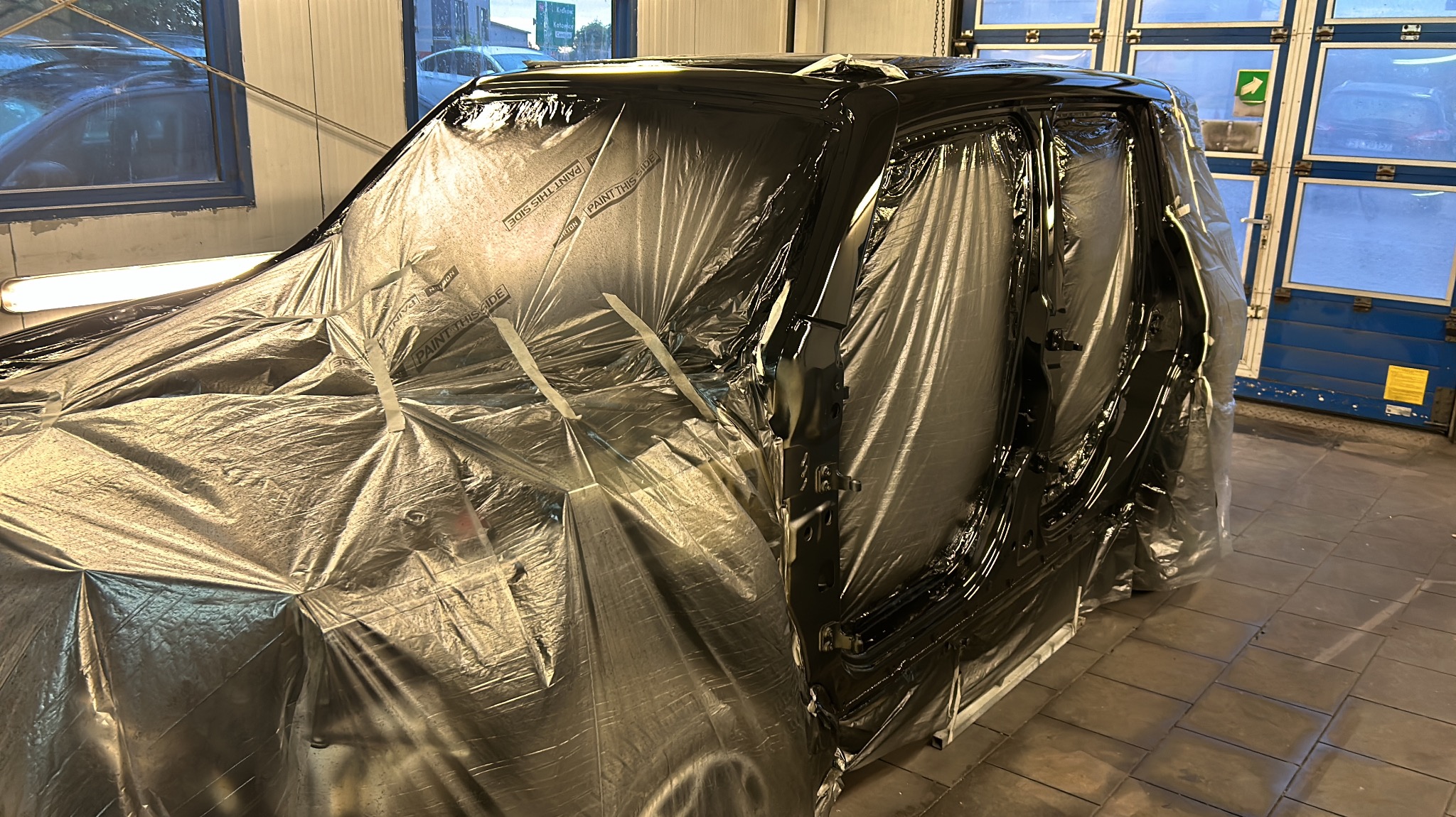 Samochód osobowy w trakcie lakierowania, zabezpieczony folią ochronną, widoczne elementy karoserii pokryte świeżą, czarną farbą.