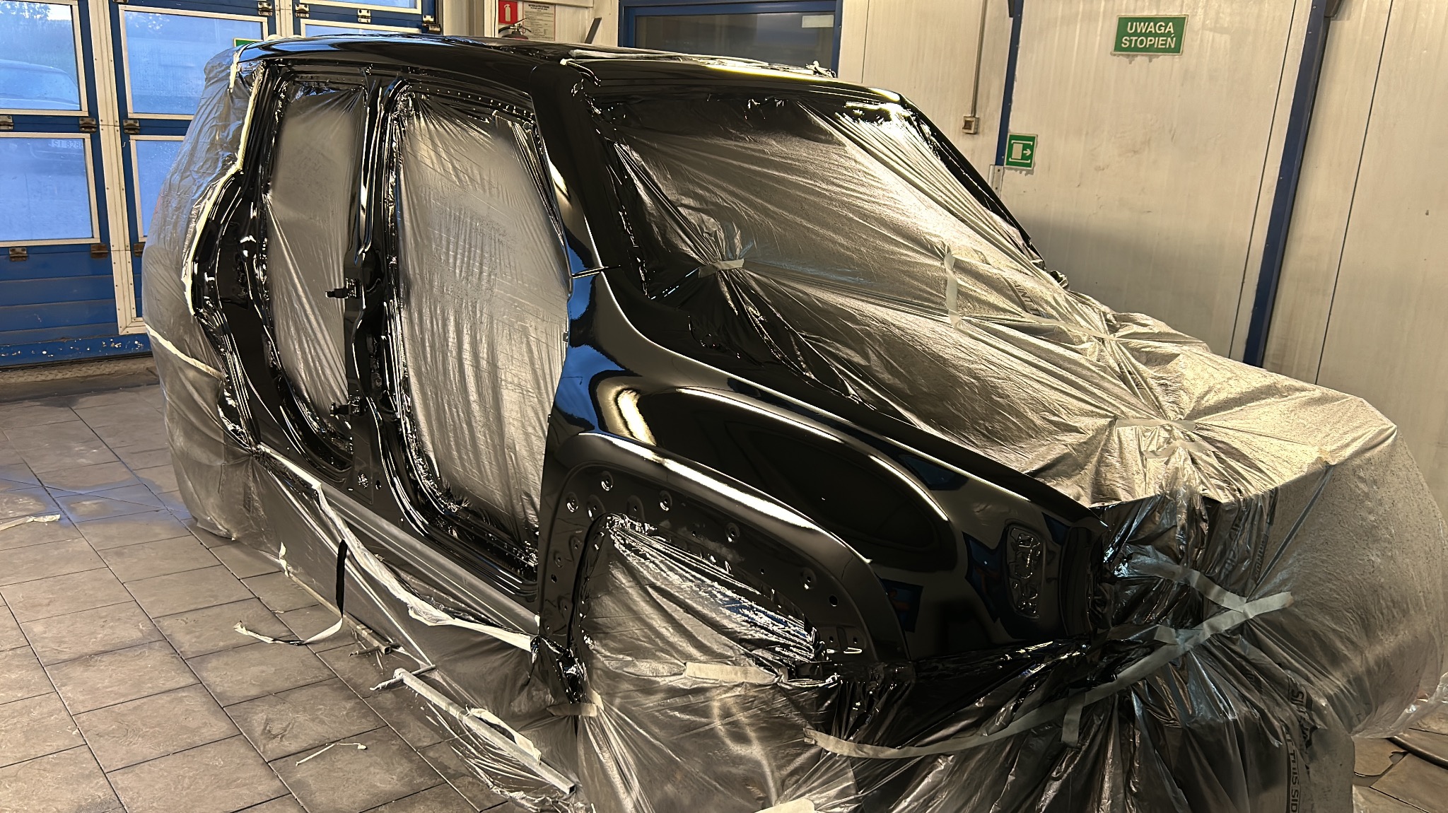 Samochód w trakcie lakierowania, zabezpieczony folią ochronną w warsztacie, widoczne świeżo polakierowane elementy karoserii.