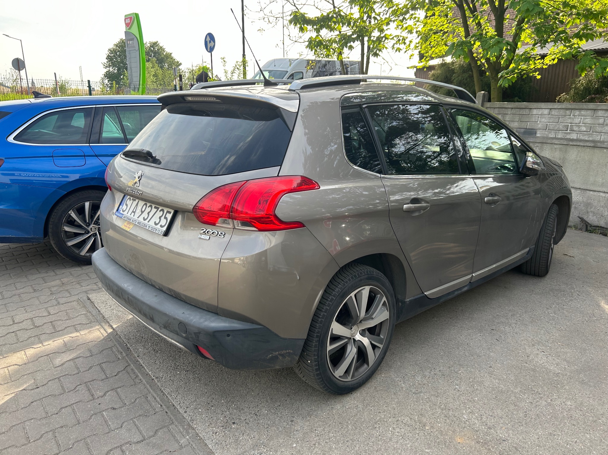 Szary Peugeot 2008 zaparkowany obok niebieskiego samochodu na chodniku, widoczny z tyłu, zarysowania na zderzaku.