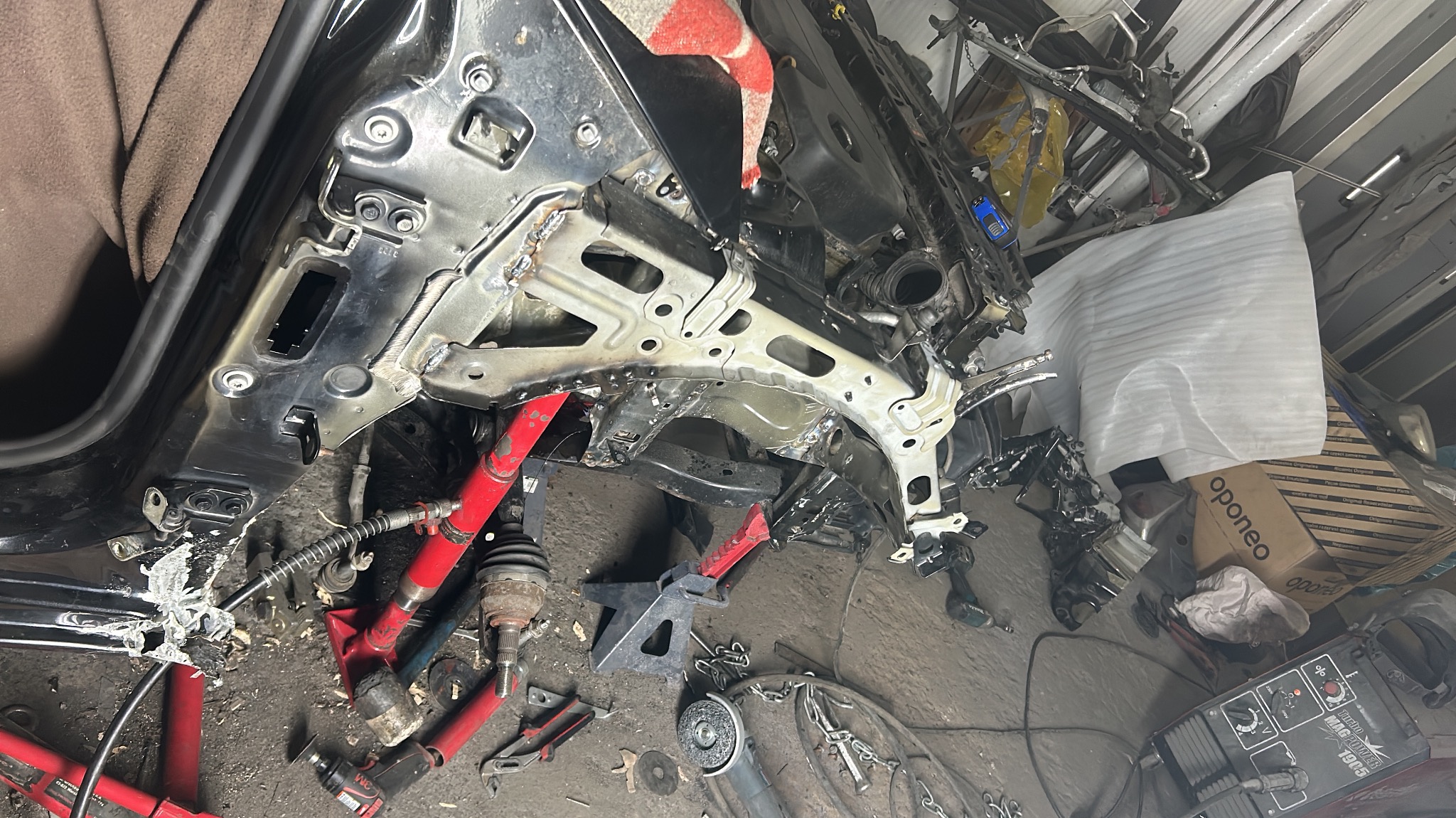 Uszkodzony fragment karoserii samochodu po spawaniu, widoczne narzędzia, podnośniki i części zamienne w warsztacie.