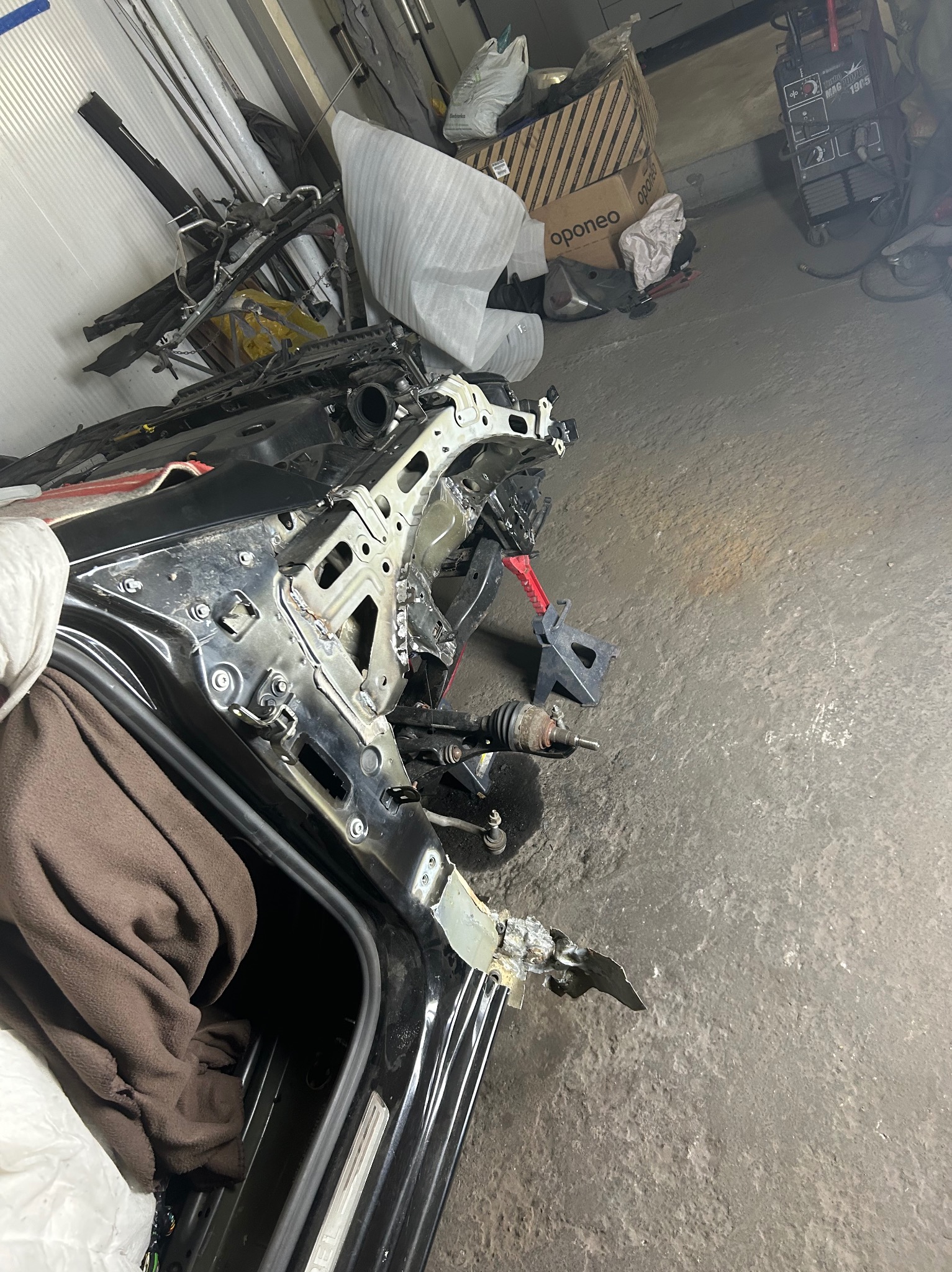 Uszkodzony przód czarnego samochodu marki Opel po wypadku, wsparty na podnośniku warsztatowym, widoczne elementy blacharskie w trakcie naprawy w warsztacie.