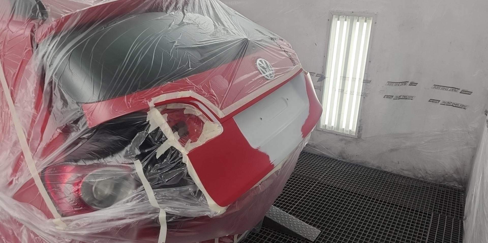 Czerwony samochód marki Volkswagen, częściowo zakryty folią ochronną i taśmą malarską, przygotowywany do lakierowania w kabinie lakierniczej z kratą podłogową.