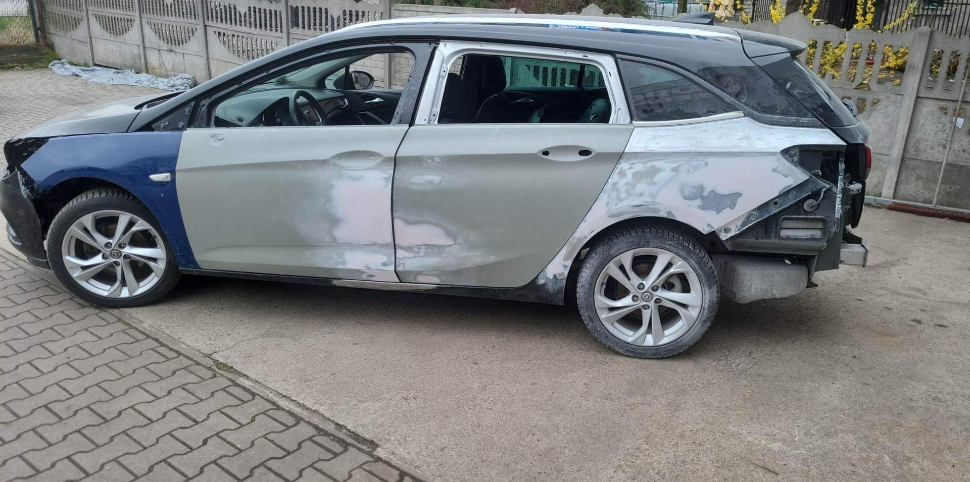 Samochód kombi w trakcie naprawy blacharskiej: widoczne fragmenty karoserii pokryte szpachlą, brak drzwi i elementów poszycia, przygotowanie do lakierowania.