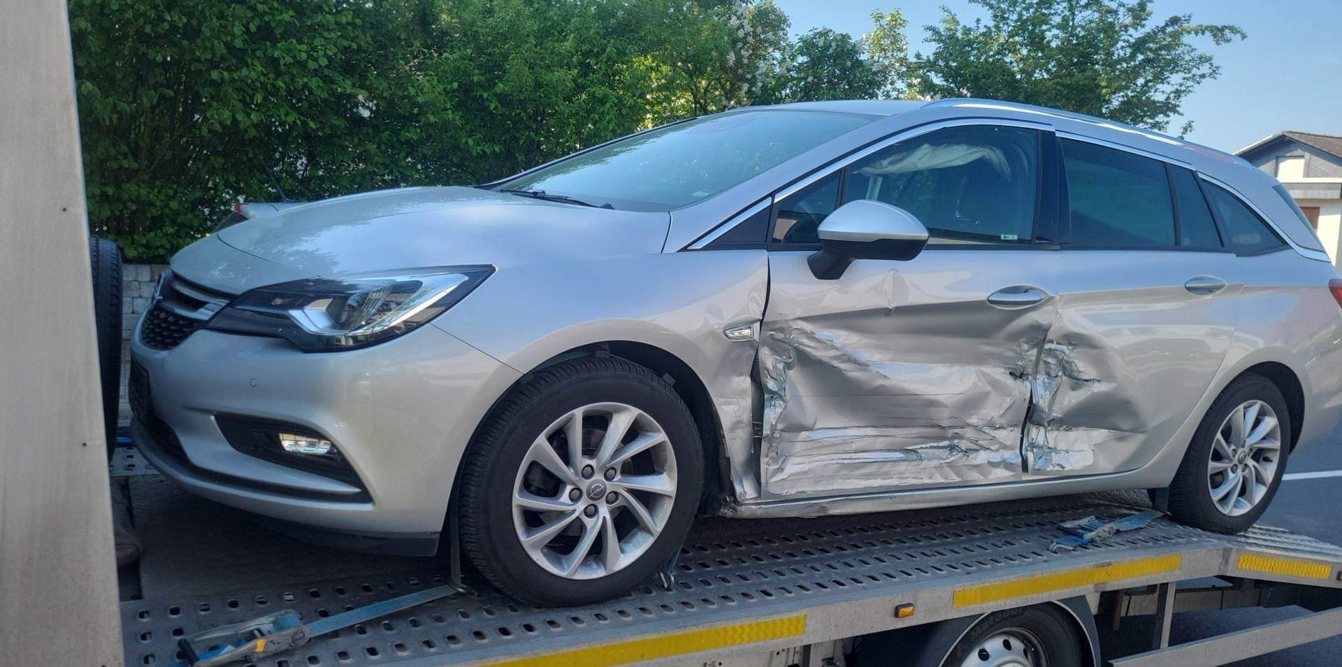 Srebrny samochód kombi z uszkodzonymi drzwiami i błotnikiem, załadowany na lawetę, widoczne zarysowania i wgniecenia.