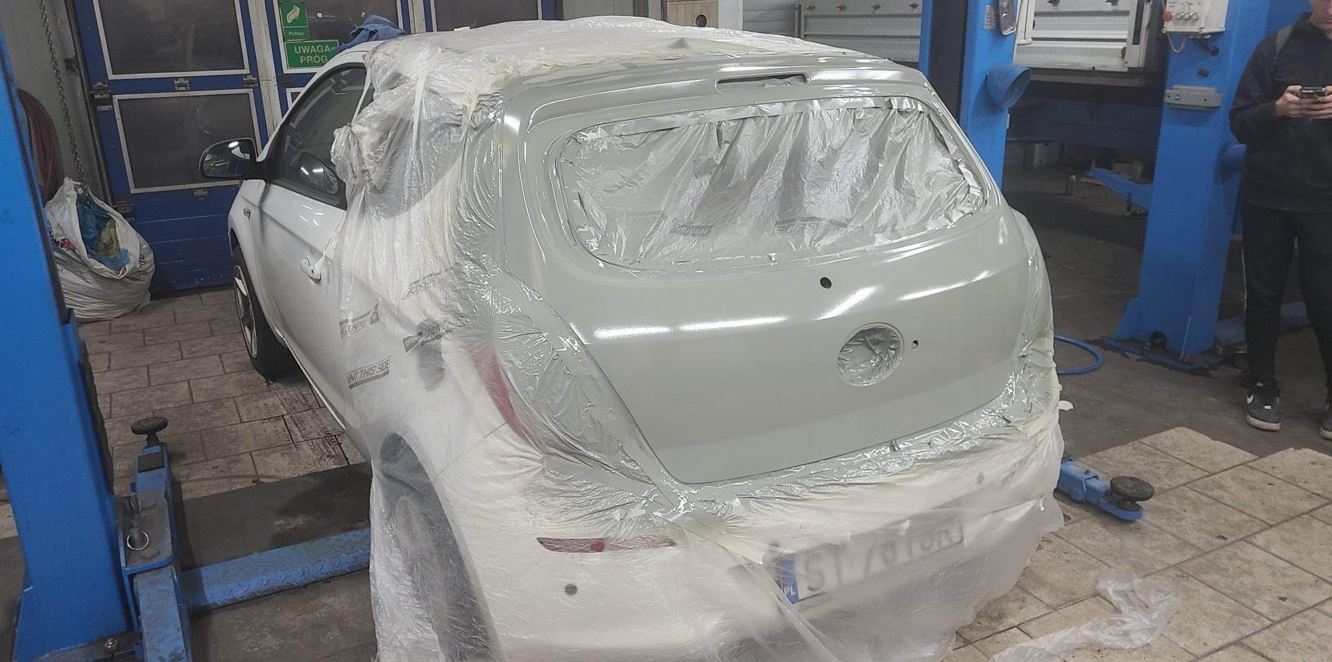 Biały samochód na podnośniku w warsztacie, częściowo oklejony folią ochronną i pokryty świeżym podkładem w kolorze szarym, przygotowany do lakierowania.