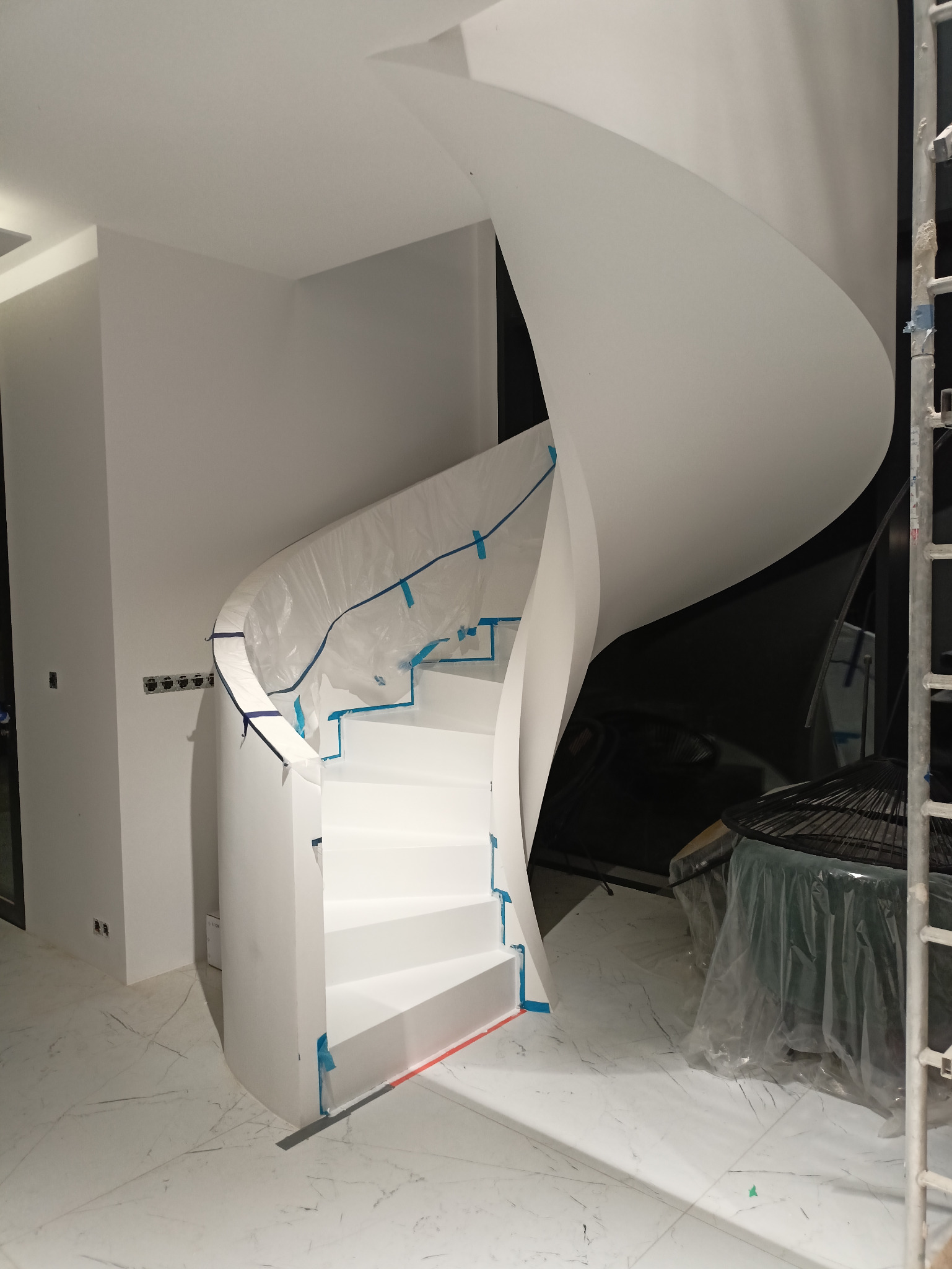 Białe, spiralne schody w trakcie malowania, zabezpieczone folią i taśmą malarską, widoczne gniazdka elektryczne na ścianie i marmurowa posadzka.