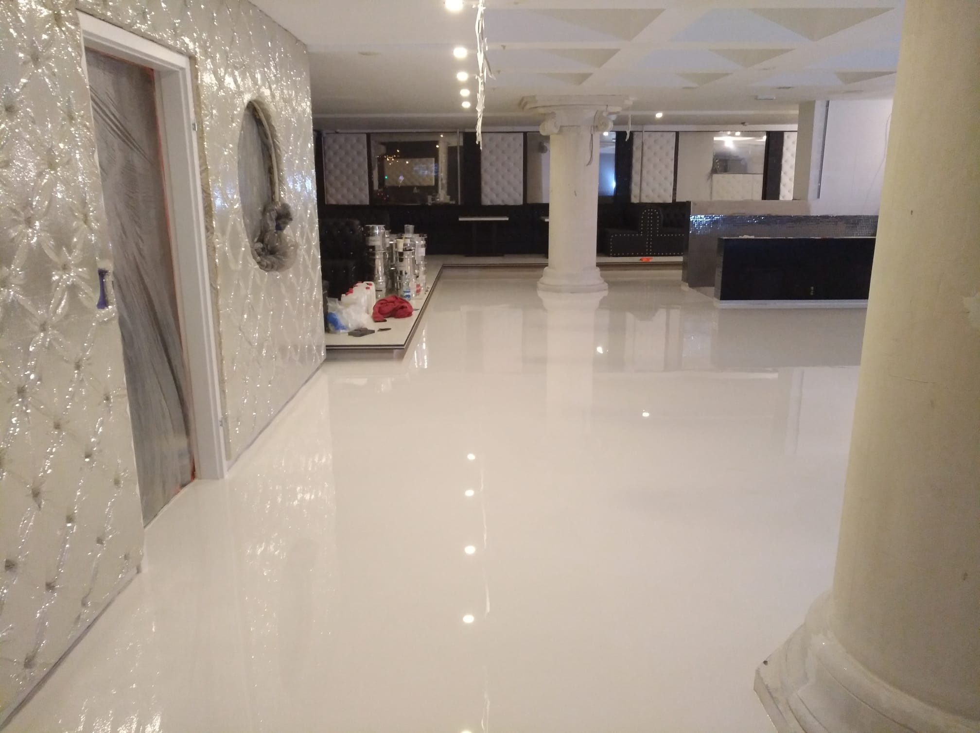 Biała, błyszcząca podłoga w przestronnym wnętrzu z kolumną, widoczne przygotowania do dalszych prac wykończeniowych.