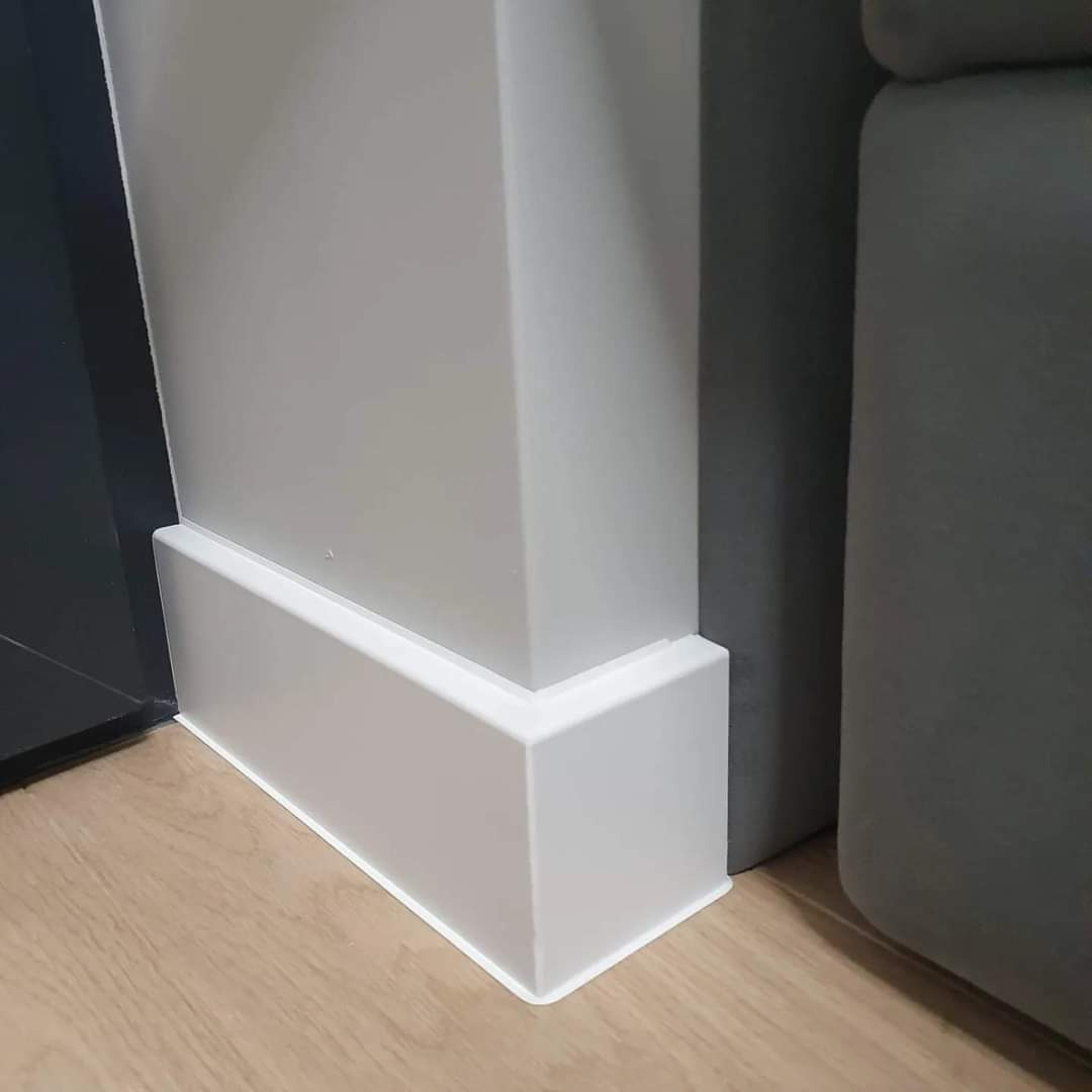 Detal wykończenia wnętrza: biała listwa przypodłogowa MDF na styku ściany, narożnika i podłogi z paneli, obok szara sofa.