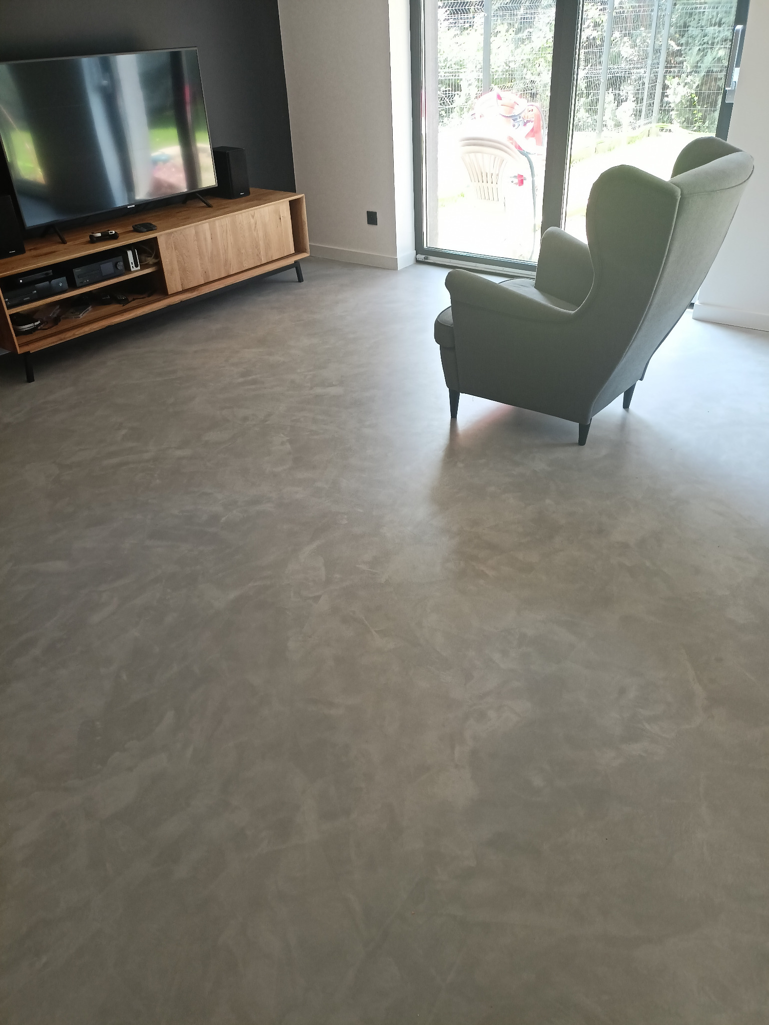 Szara, gładka podłoga z efektem chmurkowania, widoczna w salonie z drewnianą szafką RTV i fotelem typu uszak