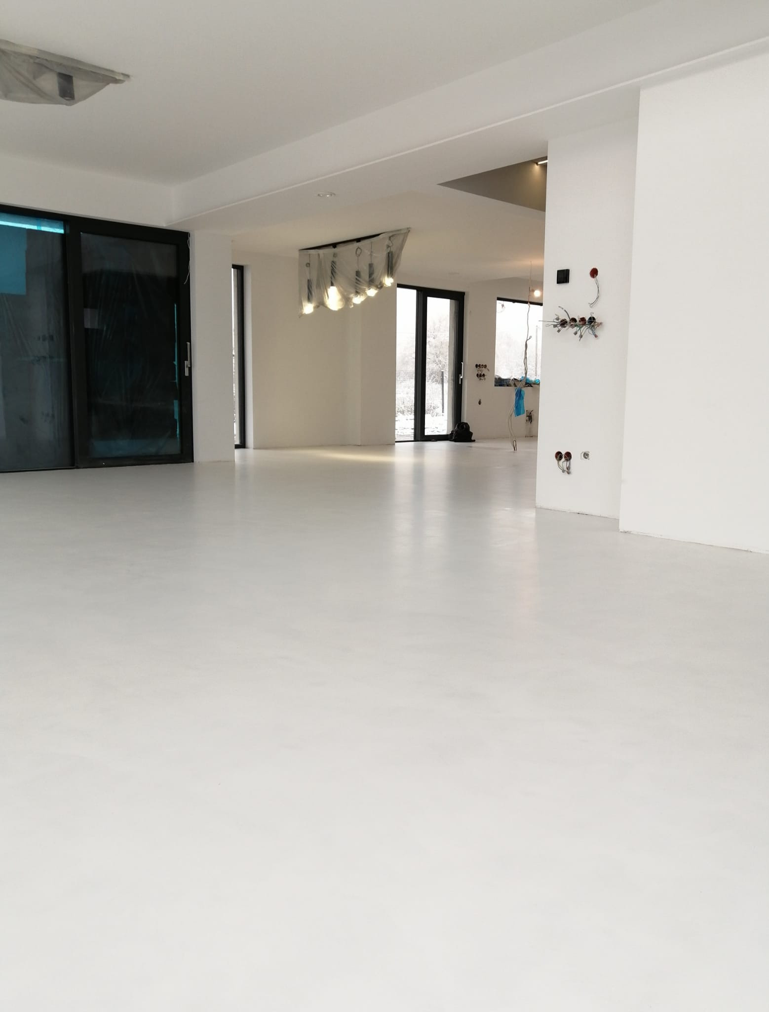 Jasne, jednolite wykończenie podłogi w przestronnym, minimalistycznym wnętrzu z dużymi oknami i przygotowanymi instalacjami elektrycznymi na ścianie.