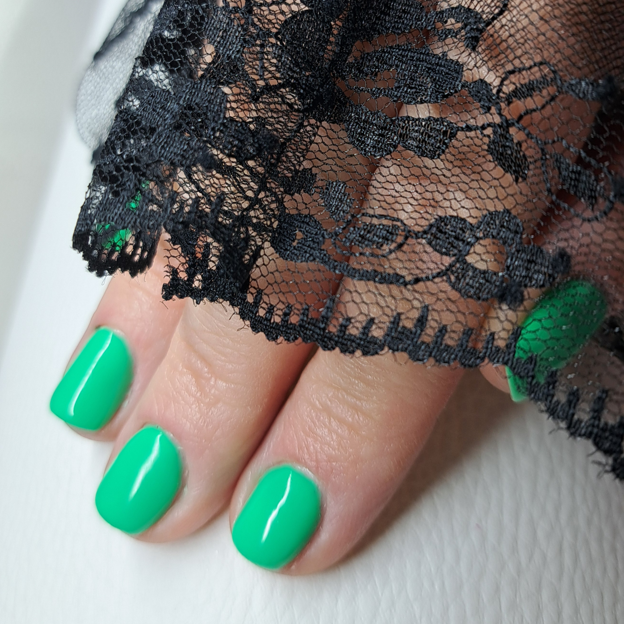 Krótkie paznokcie pomalowane na jaskrawozielony kolor, ozdobione czarną koronką na palcu wskazującym.