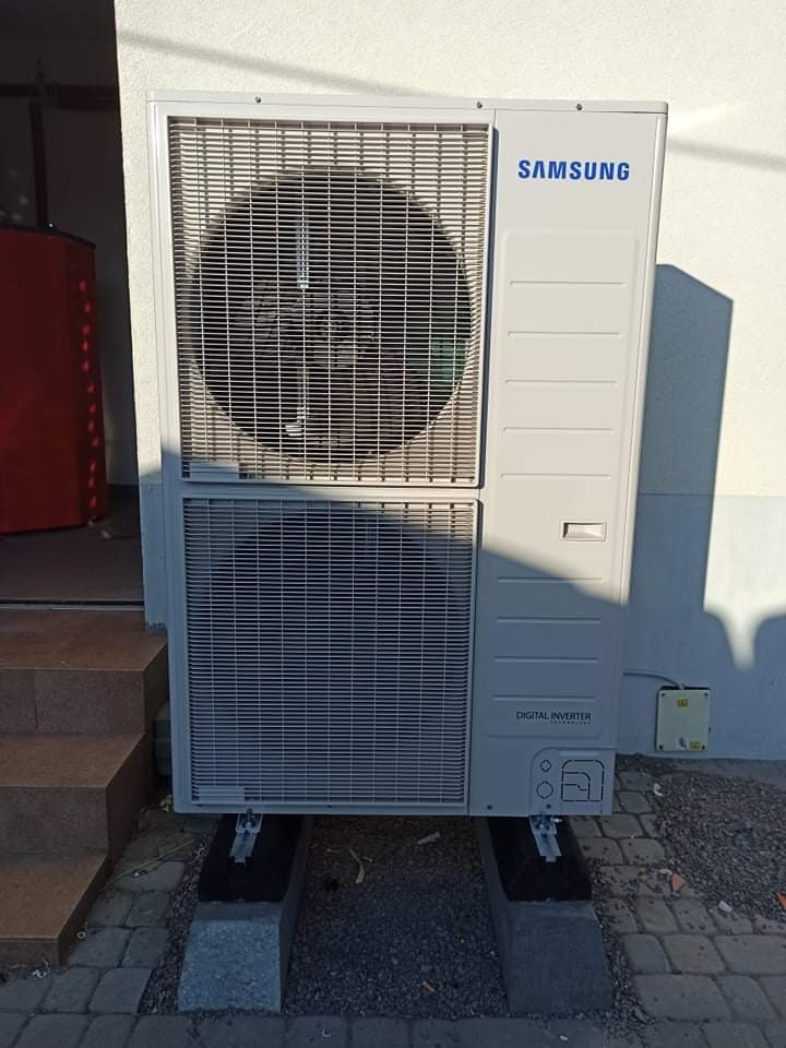 Zewnętrzna jednostka pompy ciepła marki Samsung, model Digital Inverter, zamontowana na betonowych bloczkach przy ścianie budynku i schodach wejściowych.