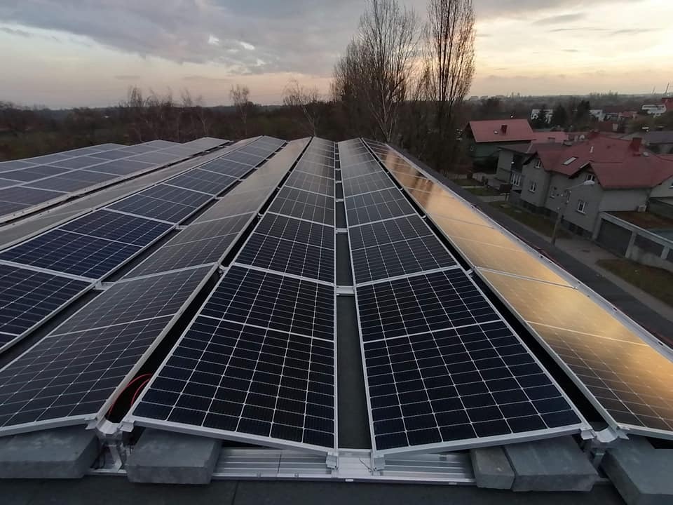 Rząd paneli słonecznych na dachu budynku, z widokiem na okoliczne domy i drzewa pod pochmurnym niebem o zachodzie słońca.