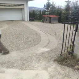 Utwardzanie podjazdu żwirowego przed garażem, widoczne ślady opon i fragment sylwetki pracownika w odzieży roboczej.