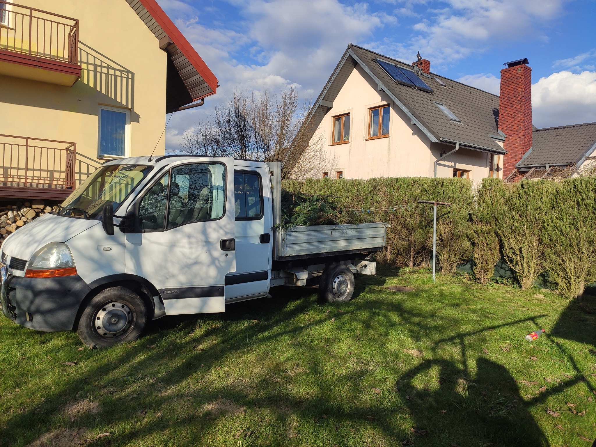 Biały samochód dostawczy z otwartą przestrzenią ładunkową wypełnioną gałęziami drzew, zaparkowany na trawniku przed domami w słoneczny dzień.