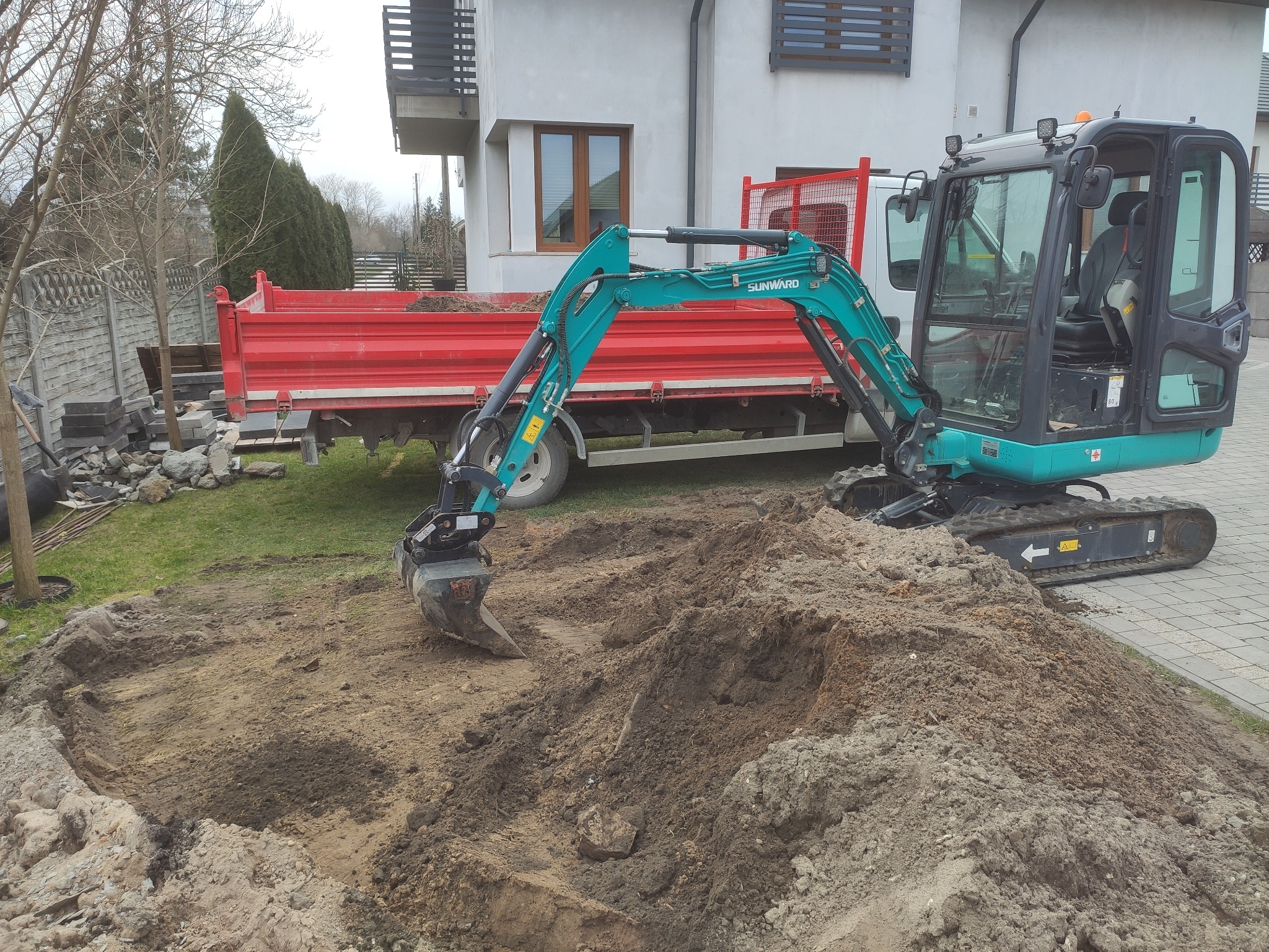 Turkusowa mini koparka Sunward na placu budowy ogrodu, z czerwoną wywrotką w tle, obok domu z szarą elewacją.