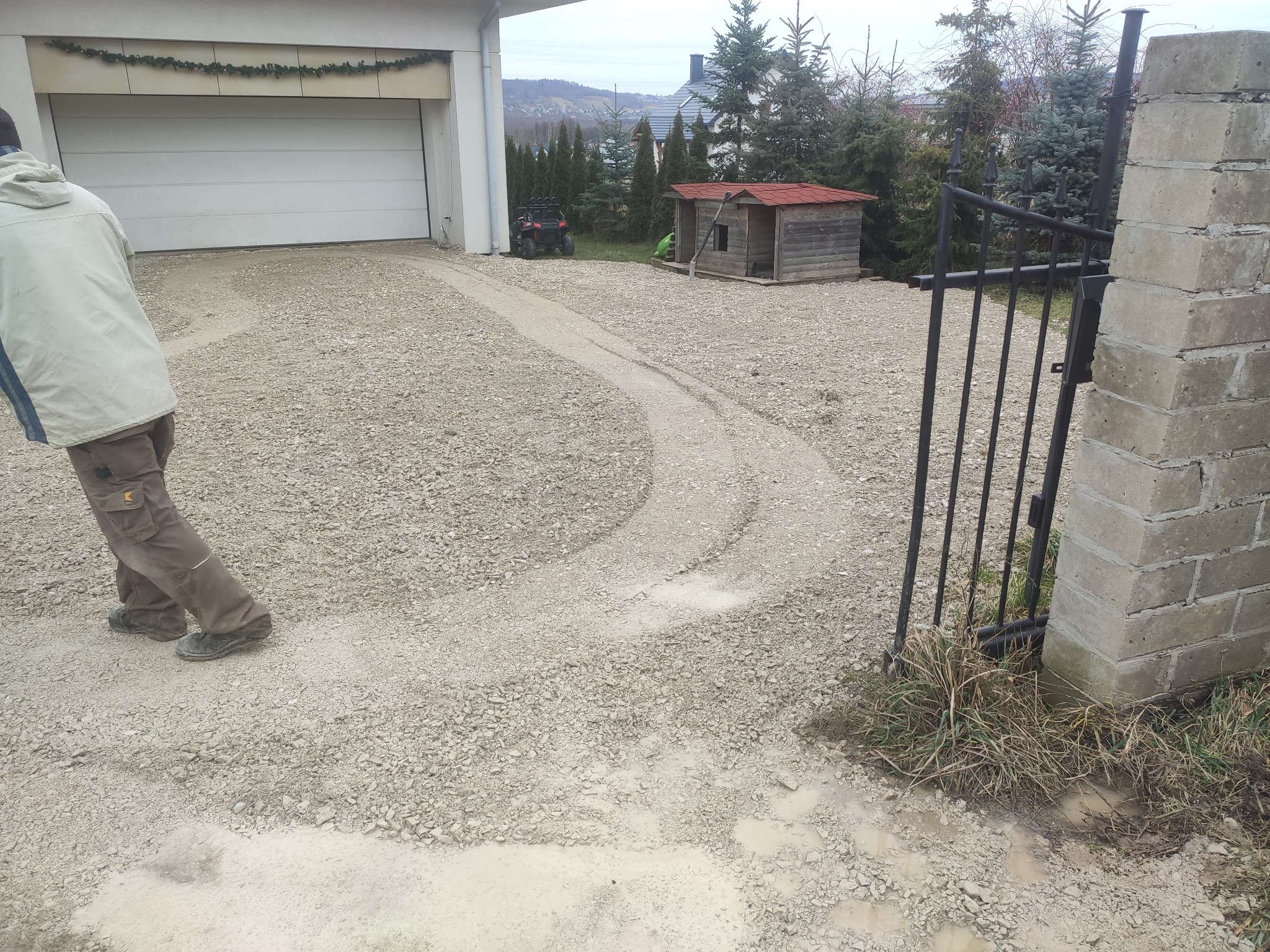 Utwardzanie podjazdu żwirowego przed garażem, widoczne ślady opon i fragment sylwetki pracownika w odzieży roboczej.