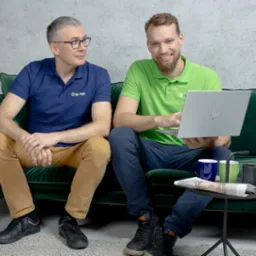 Dwóch mężczyzn siedzi na zielonej kanapie. Jeden z nich, ubrany w zieloną koszulkę polo, trzyma srebrnego laptopa i uśmiecha się. Drugi, w granatowej koszulce polo, patrzy na niego. Obaj mają logo...