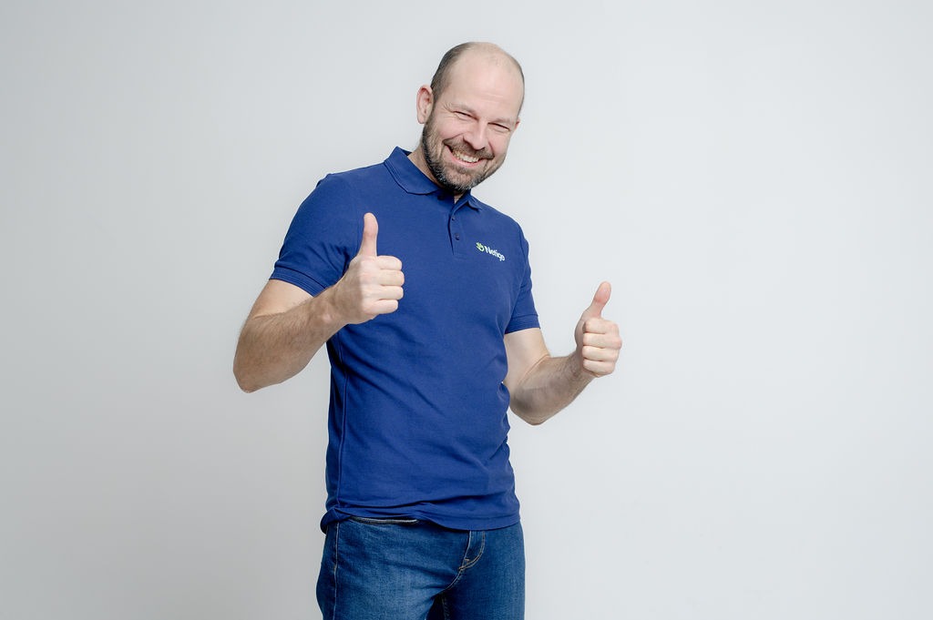 Uśmiechnięty mężczyzna w niebieskiej koszulce polo z logo, gestykulujący oboma kciukami w górę na jasnym tle.