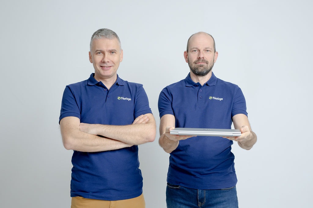 Dwóch mężczyzn w granatowych koszulkach polo z logo firmy prezentuje zamknięty laptop na białym tle.