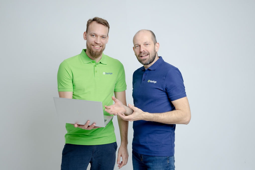 Dwóch uśmiechniętych mężczyzn w koszulkach polo z logo firmy, jeden trzyma otwarty laptop, gestykulują na jasnym tle.