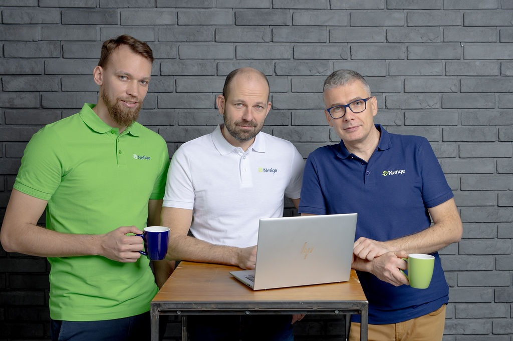 Trzech mężczyzn w koszulkach polo z logo firmy Netige stoi za drewnianym stołem z laptopem HP, trzymając kubki na tle szarej ściany z cegieł.