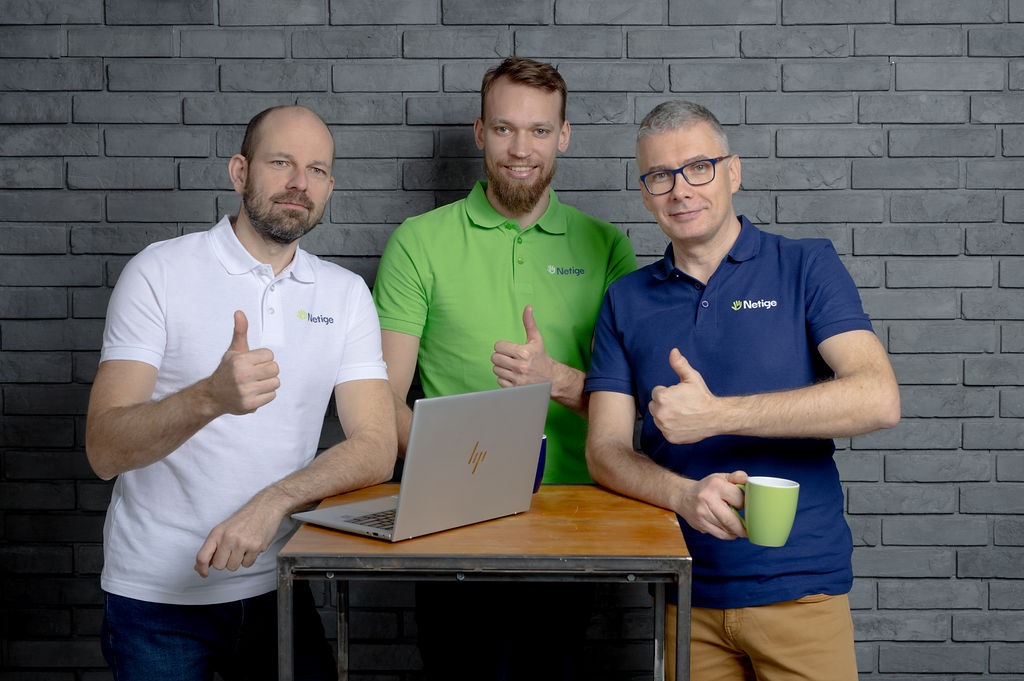 Trzech uśmiechniętych mężczyzn w koszulkach polo z logo firmy, stojących za stołem z laptopem, pokazujących kciuki w górę na tle szarej ściany z cegieł.