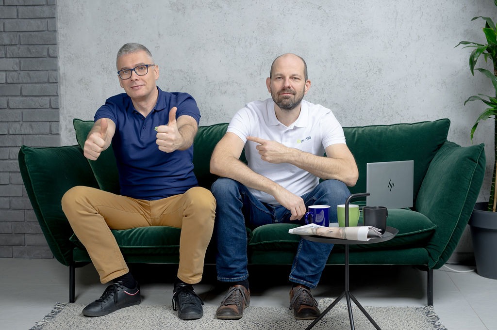 Dwaj mężczyźni siedzą na szmaragdowej sofie, jeden w okularach pokazuje dwa kciuki w górę, drugi wskazuje palcem, obok stoi stolik z laptopem, kubkami i gazetą.