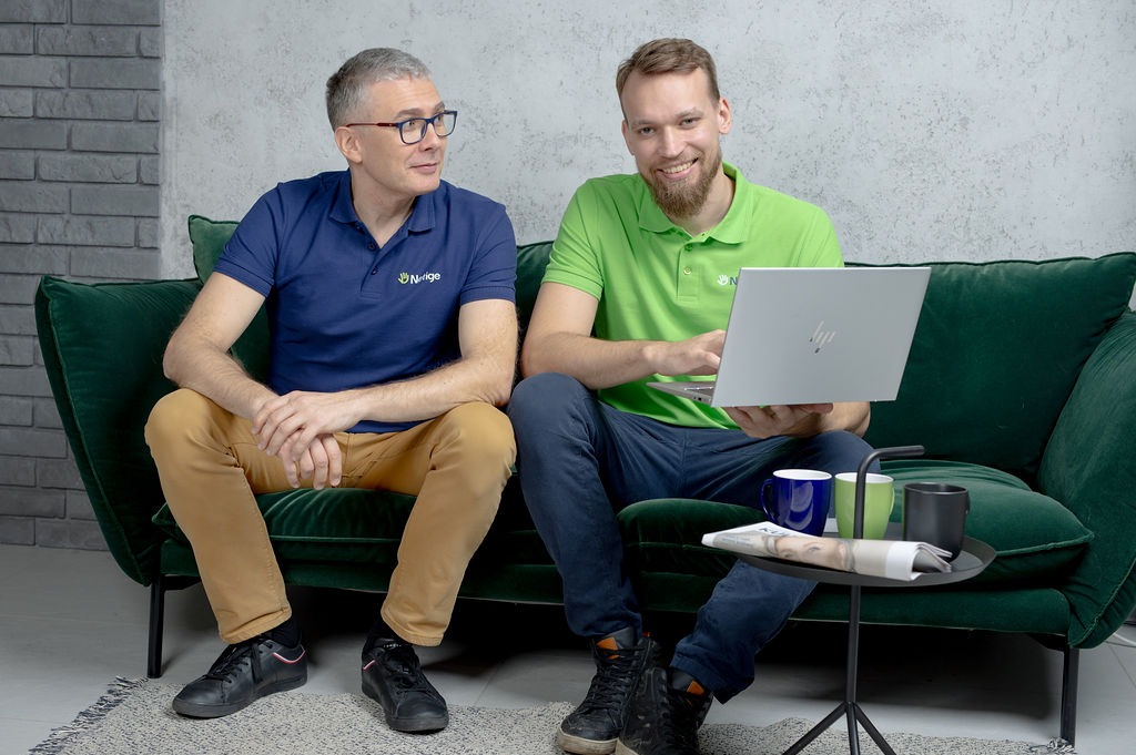 Dwóch mężczyzn siedzi na zielonej kanapie. Jeden z nich, ubrany w zieloną koszulkę polo, trzyma srebrnego laptopa i uśmiecha się. Drugi, w granatowej koszulce polo, patrzy na niego. Obaj mają logo...