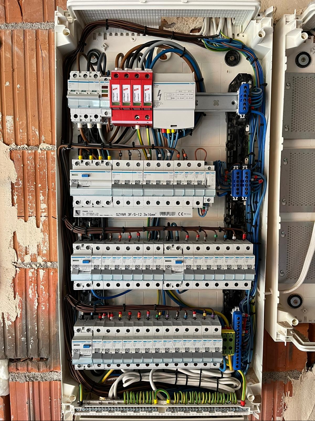 Rozdzielnica elektryczna z widocznymi zabezpieczeniami przepięciowymi (czerwone moduły), bezpiecznikami automatycznymi marki Hager, listwami zaciskowymi i okablowaniem, zamontowana na ścianie z cegły.