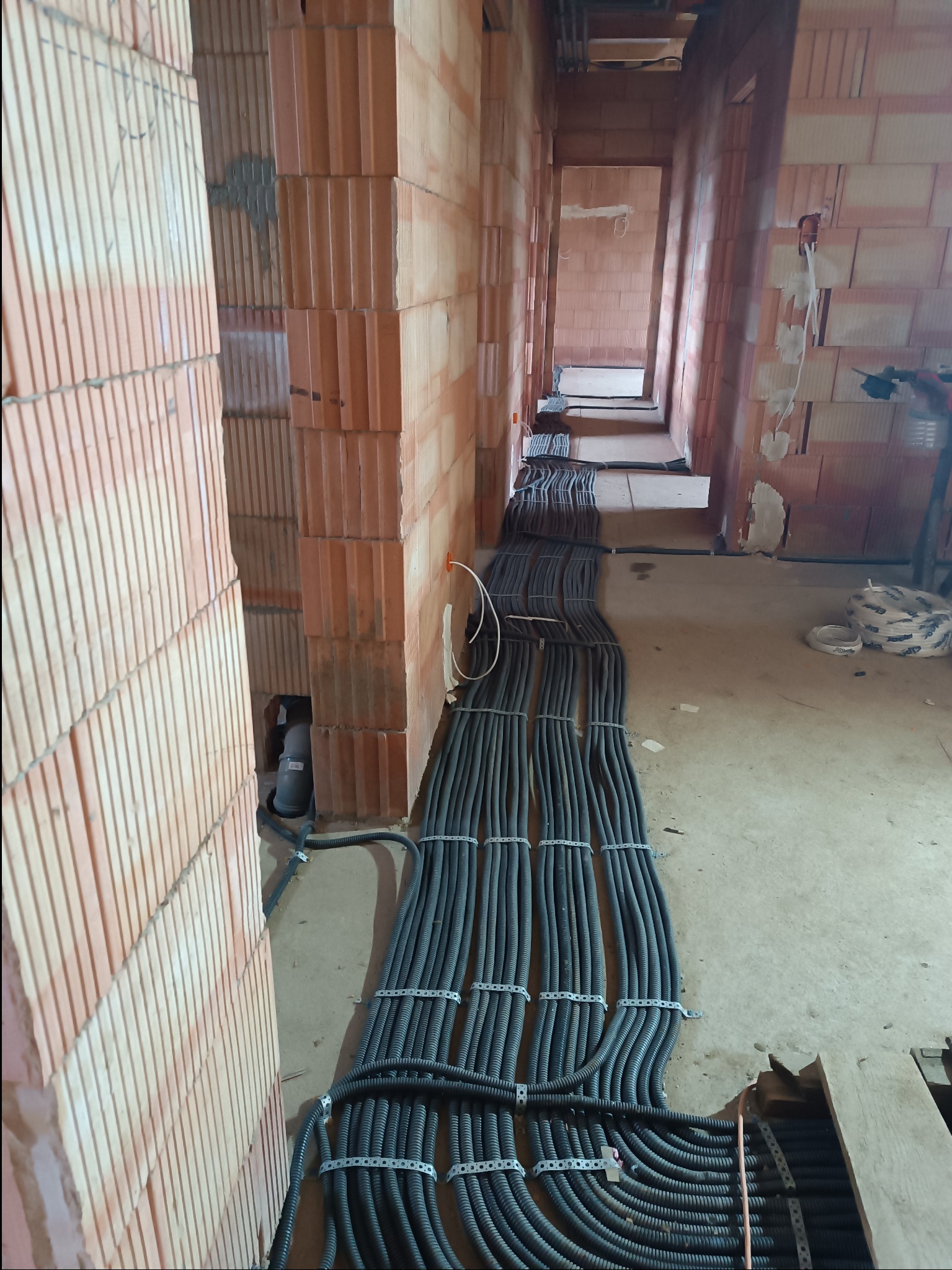 Równolegle ułożone, czarne rury karbowane z kablami, mocowane do podłoża metalowymi uchwytami, prowadzone wzdłuż surowych ścian z czerwonej cegły dziurawki w budynku w trakcie budowy.