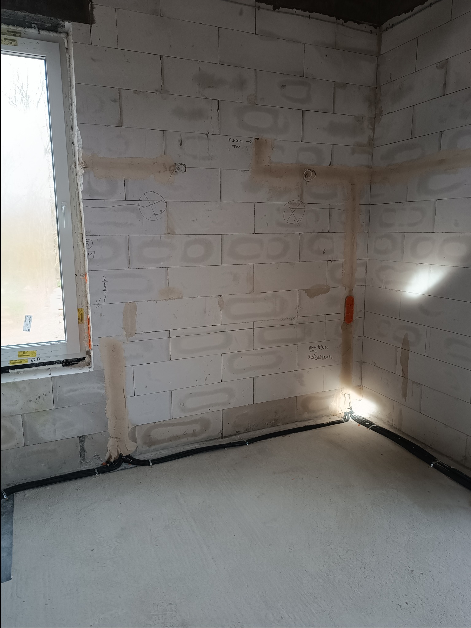 Wnętrze w trakcie prac instalacyjnych: surowe ściany z betonu komórkowego z widocznymi trasami kablowymi, puszkami pod gniazdka i włączniki, oraz czarnymi rurkami karbowanymi przy podłodze, z oknem...