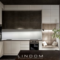 LINDOM ARCHITEKTURA SPÓŁKA Z OGRANICZONĄ ODPOWIEDZIALNOŚCIĄ - Nowoczesna kuchnia z białymi szafkami, ciemnymi płytkami nad blatem i wnęką z siedziskiem obok okna, oświetlona lampką punktową i dekorowana gałązką oliwną w szklanym wazonie.