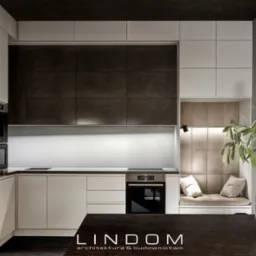 Nowoczesna kuchnia z białymi szafkami, ciemnymi płytkami nad blatem i wnęką z siedziskiem obok okna, oświetlona lampką punktową i dekorowana gałązką oliwną w szklanym wazonie.