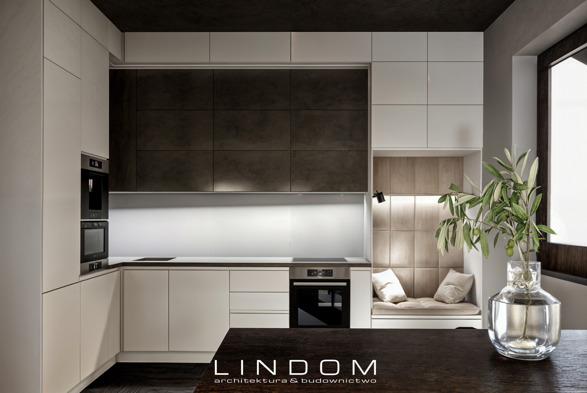 Nowoczesna kuchnia z białymi szafkami, ciemnymi płytkami nad blatem i wnęką z siedziskiem obok okna, oświetlona lampką punktową i dekorowana gałązką oliwną w szklanym wazonie.