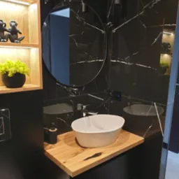 Nowoczesna łazienka z umywalką nablatową na drewnianym blacie, czarne marmurowe ściany z okrągłym lustrem i dekoracyjnymi figurkami małp w podświetlanej wnęce.
